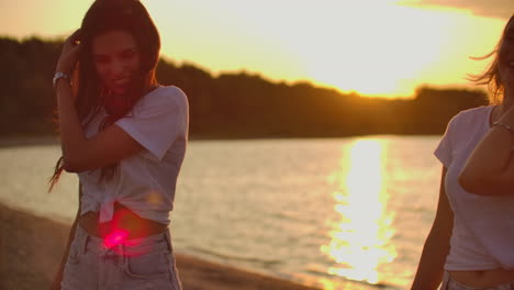 Two-young-girls-in-white-T-shirts-are-enjoying-summer-open-air-party-on-the-beach.-They-smile-and-touch-their-long-hair-at-sunset.